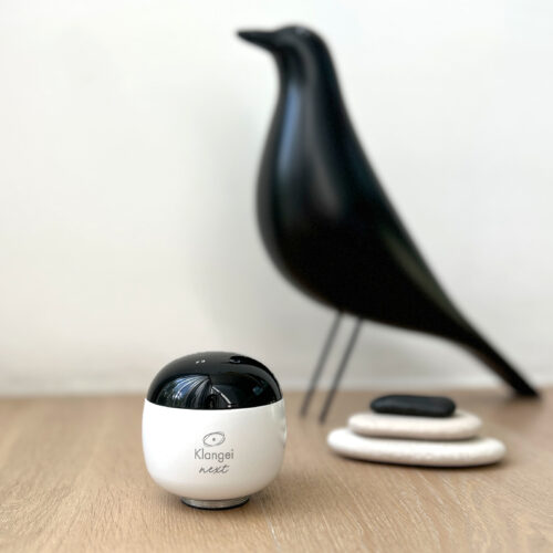 White cylindrical microphone with a black top on a wooden desk, logo near the bottom, in a blurred office setting.