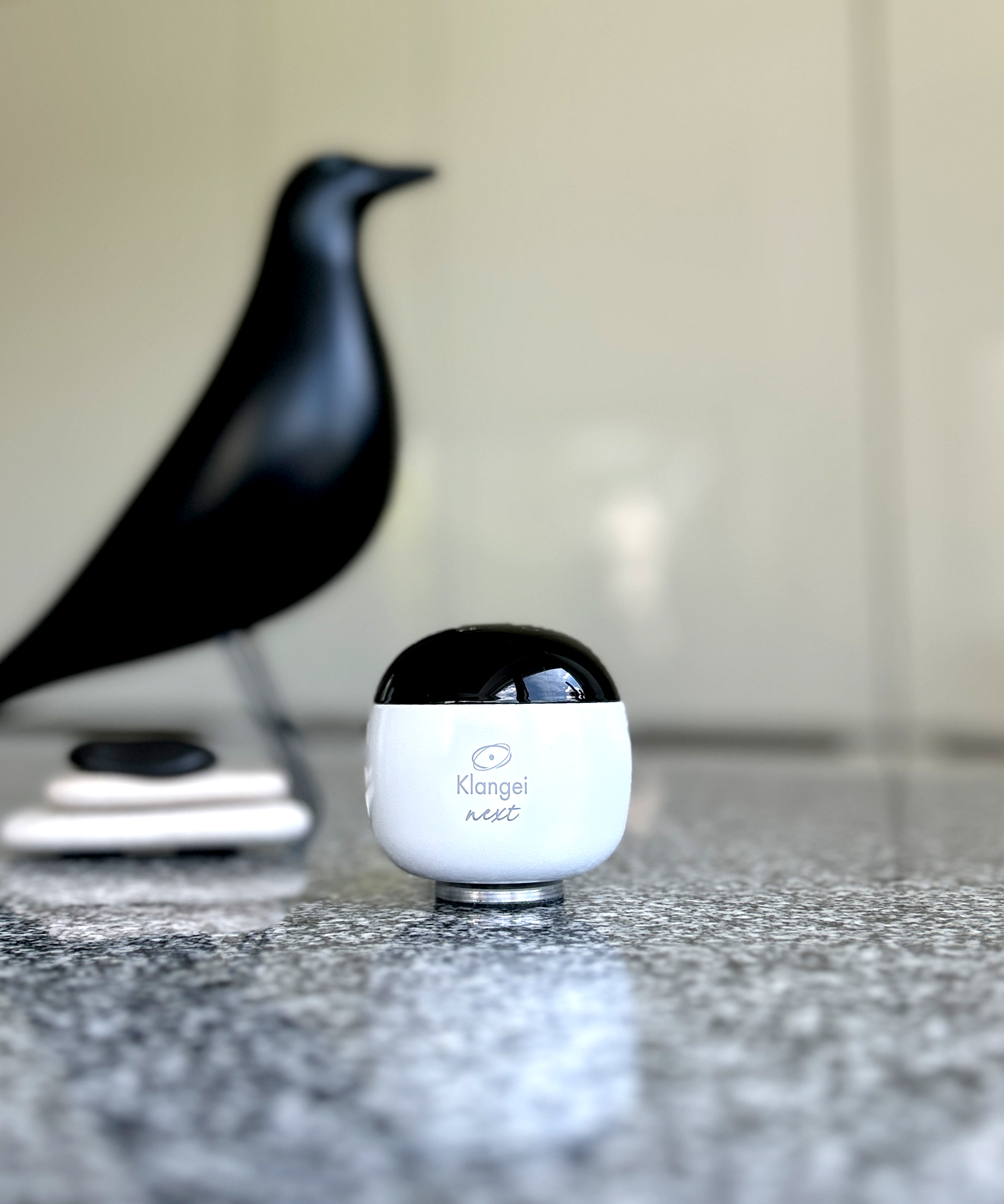 White rounded device with a black top on a speckled kitchen counter; blurred black objects in the background.