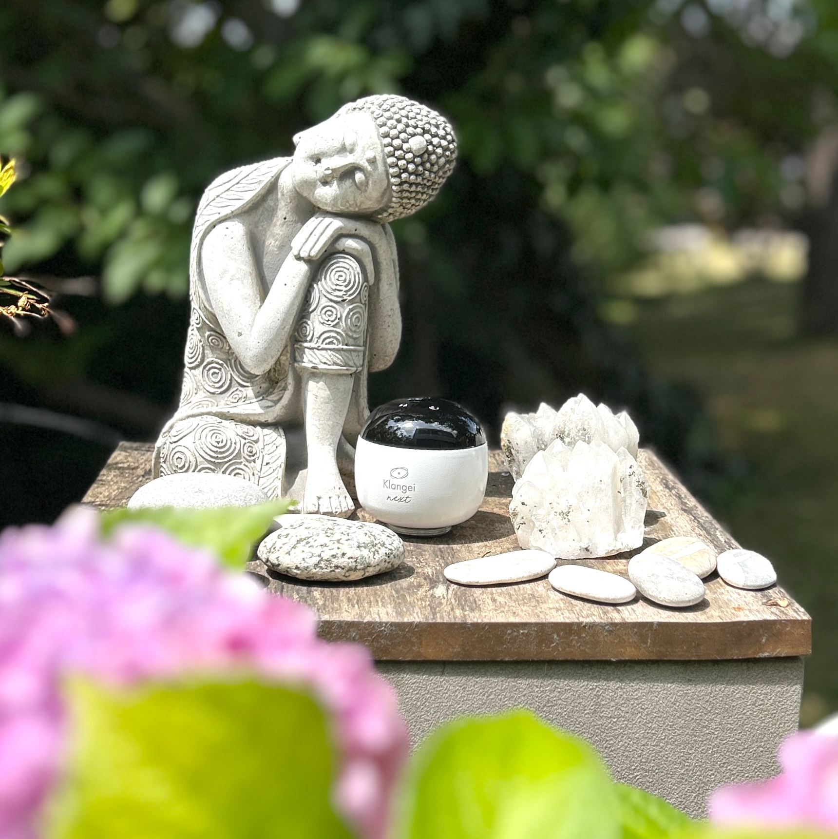 Gray seated statue with head resting on its knee on a wooden pedestal, surrounded by smooth stones and crystals in a garden setting.