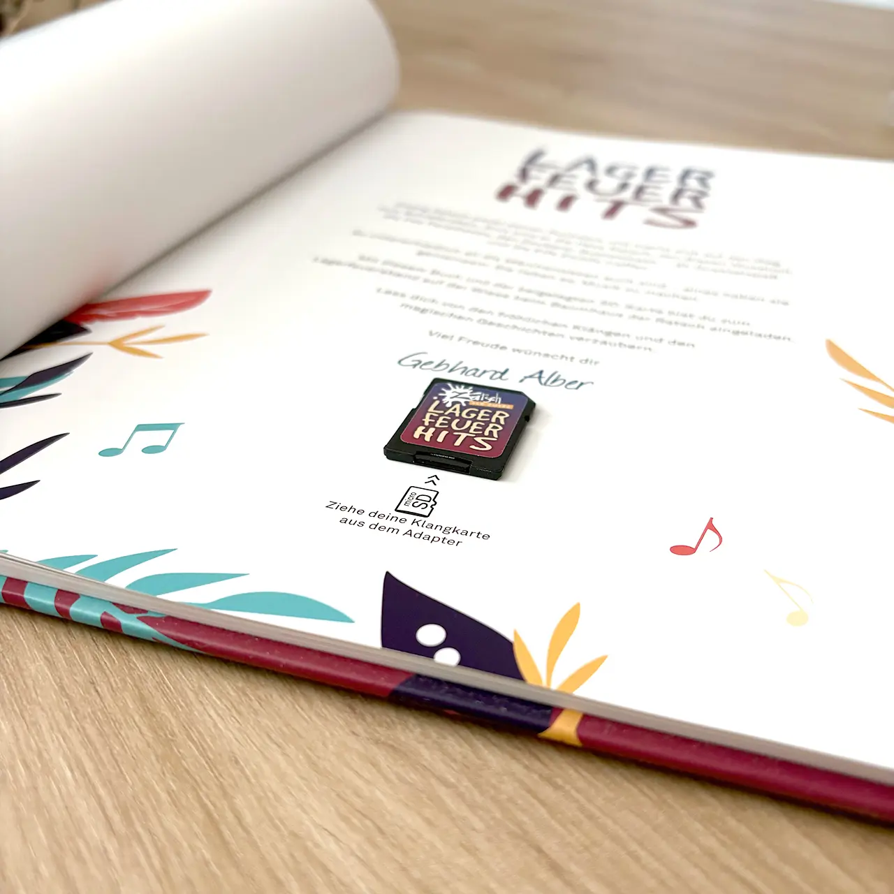 Open book page with colorful decorative margins and a small SD card labeled 'Lagerfeuer Hits' resting on the page.