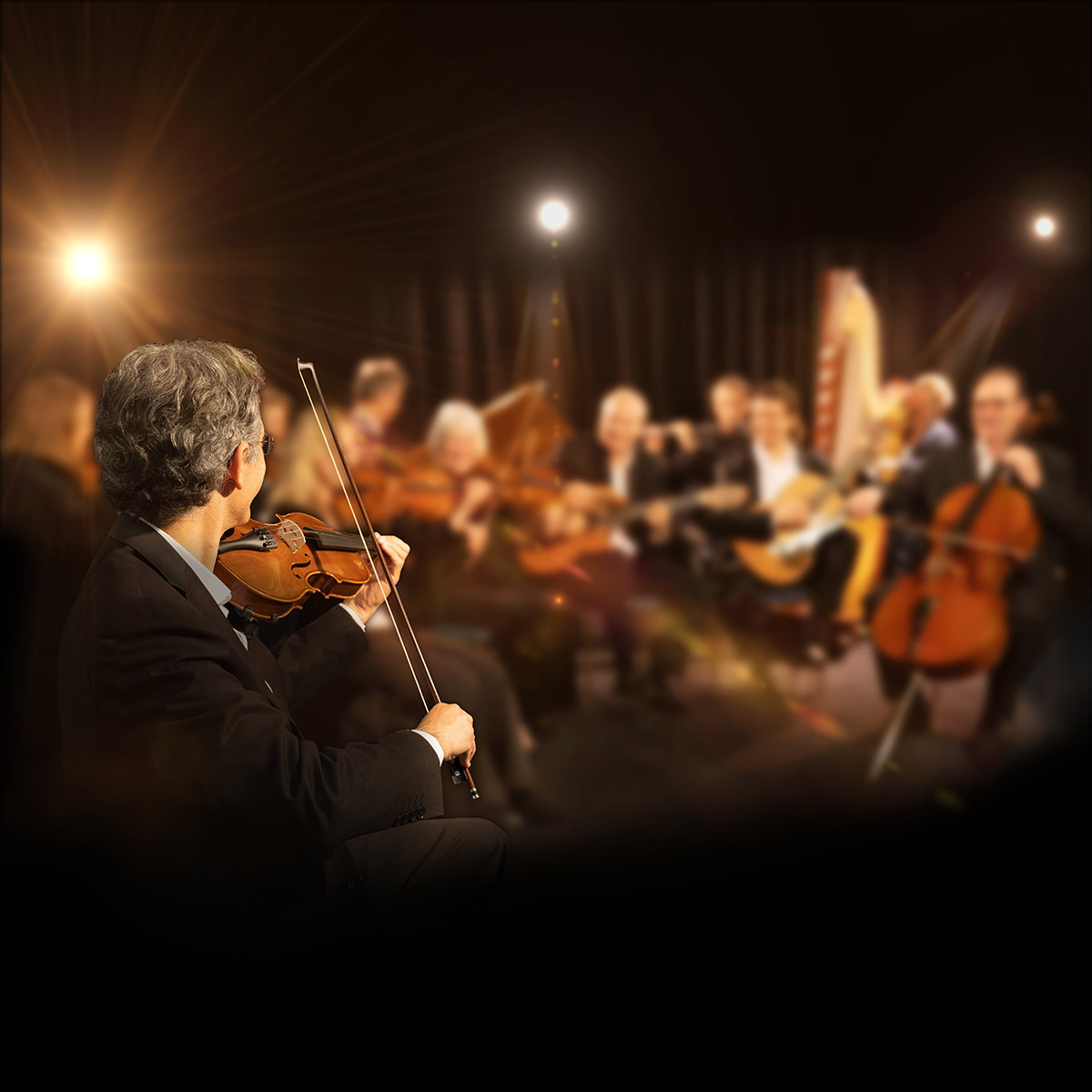 Violinist in the foreground plays on stage with a blurred orchestra behind him under warm stage lights.